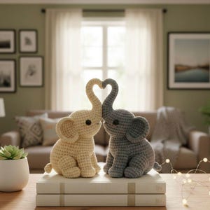 以下が含まれることがあります： クリーム色とグレーの2つの編み象の置物が並んでおり、鼻がハート型になっています。象は白い本の上に置かれ、背景にはニュートラルな色調のリビングルームがあります。