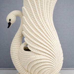 Op de afbeelding: Een crèmekleurige, gehaakte zwaan sculptuur. De zwaan heeft een lange nek en een zwarte snavel. De vleugels zijn uitgespreid en de sculptuur heeft een gestructureerd, geribbeld ontwerp. De zwaan is een decoratief item.