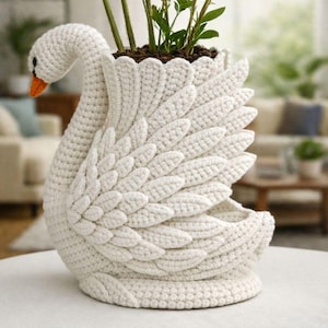 Pode incluir: Um vaso de flores de cisne de crochê branco sujo. O cisne apresenta asas detalhadas, um pescoço curvo e um bico laranja. Contém terra e plantas verdes, ideal para decoração.