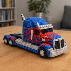 May include: A handmade crochet toy truck in blue, red, and grey. The truck features a blue cab with red flame details, a grey chassis, and black wheels. The toy is a detailed replica of a semi-truck.