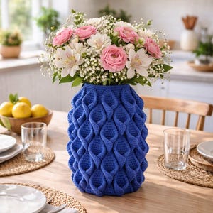 Puede incluir: Un jarrón azul de ganchillo con un diseño texturizado contiene un ramo de rosas rosas y flores blancas. El jarrón está sobre una mesa de madera, con cubiertos y vasos.