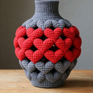 Puede incluir: Un jarrón de ganchillo gris con un patrón de corazones rojos. El jarrón hecho a mano tiene una apariencia texturizada. Los corazones están espaciados uniformemente y añaden un toque decorativo. Un objeto decorativo.