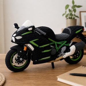 Może przedstawiać: Ręcznie wykonany model motocykla z czarnej i zielonej włóczki. Zabawkowy motocykl ma białe akcenty, zielone detale i czarne siedzenie. Koła mają zielone felgi. Model jest wystawiony na drewnianej powierzchni.