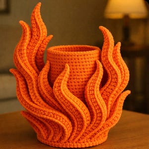 May include: An orange crochet vase with flame-like accents. The handmade vase features a cylindrical center surrounded by upward-curving, flame-shaped elements, creating a unique and decorative piece. The yarn is a solid, vibrant orange.