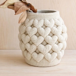 Op de afbeelding: Crèmekleurige, handgemaakte vaas met een hartvormig patroon. De vaas is gemaakt van een getextureerd, geweven materiaal en bevat gedroogde bladeren. Een decoratief stuk voor de woondecoratie, ongeveer 20 cm hoog.