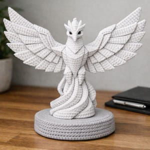 Op de afbeelding: Een gehaakte witte fenikssculptuur met uitgespreide vleugels, geplaatst op een grijze cirkelvormige basis. De gedetailleerde veren en kop van de vogel zijn zorgvuldig vervaardigd, wat een getextureerde, handgemaakte uitstraling laat zien. De sculptuur staat op een houten oppervlak.