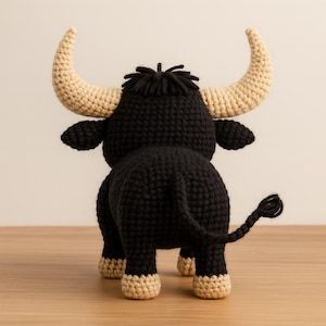 May include: A crocheted bull toy, seen from behind. The bull is mainly black with beige horns, hooves, and details. The toy has a textured finish and is placed on a wooden surface.