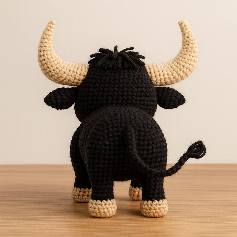 May include: A crocheted bull toy, seen from behind. The bull is mainly black with beige horns, hooves, and details. The toy has a textured finish and is placed on a wooden surface.