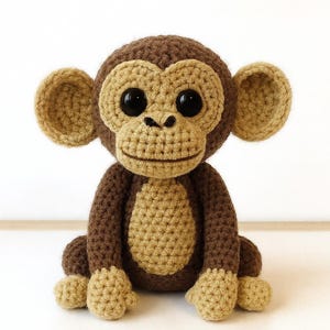 Puede incluir: Una muñeca de mono de ganchillo hecha a mano en tonos marrón y beige. El mono tiene grandes ojos negros, una cara, orejas, manos y pies beige y un cuerpo marrón. El mono amigurumi está sentado erguido.