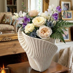 Könnte beinhalten: Eine cremefarbene, gestrickte Vase enthält einen bunten Blumenstrauß. Das Arrangement umfasst hellgelbe Rosen, lila Platterbsen und blaue Disteln. Die Vase hat ein einzigartiges, strukturiertes Design und steht auf einem Holztisch.