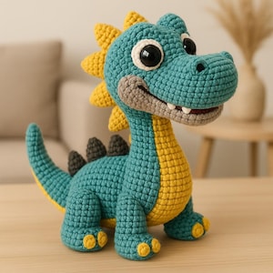 Puede incluir: Un juguete de dinosaurio tejido a ganchillo en verde azulado, amarillo y gris. El peluche hecho a mano tiene grandes ojos negros, una boca sonriente y púas amarillas a lo largo de la espalda. El dinosaurio es un diseño lindo y amigable.