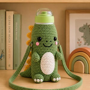 May include: A green crochet water bottle holder shaped like a dinosaur with a yellow spiked back, a white belly patch, and two small arms. The holder features a long green crochet strap, black eyes, and pink cheeks, topped with a light green plastic lid.