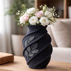 Puede incluir: Un jarrón de ganchillo azul marino con un diseño en espiral y detalles de hojas bordadas. El jarrón contiene un ramo de rosas blancas y rosas y pequeñas flores blancas. El jarrón está sobre una mesa de madera.