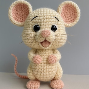 May include: A crocheted mouse toy in cream with pink accents. The mouse has large black eyes, a pink nose, and pink inner ears. Its paws and tail are also pink. The mouse is smiling and appears to be handmade.