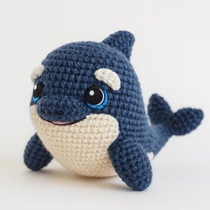 May include: A handmade crochet orca whale toy. The orca is primarily dark blue with a cream-colored belly and white accents. It has black and blue eyes and a smiling mouth. The toy is designed to look like a friendly cartoon character.