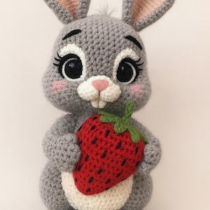 May include: A handmade gray crochet bunny holding a red strawberry. The bunny has large black eyes, pink cheeks, and long ears with pink inner ears. The strawberry has green leaves and black seeds.