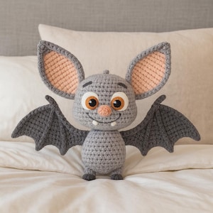 Pode incluir: Uma boneca de morcego de crochê com uma expressão amigável. O morcego é principalmente cinza, com asas e orelhas grandes e texturizadas. O interior das orelhas é cor de pêssego, e os olhos são grandes com detalhes em laranja e branco. Tem um pequeno nariz rosa e uma boca sorridente.