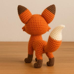 K&ouml;nnte beinhalten: Eine handgefertigte Fuchsfigur aus H&auml;kelarbeit. Der Fuchs ist haupts&auml;chlich orange mit braunen Akzenten an Ohren und F&uuml;&szlig;en. Der Schwanz ist wei&szlig; mit einem orangefarbenen Ansatz. Der Fuchs steht auf einer Holzoberfl&auml;che. Ca. 15 cm hoch.