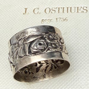 Könnte beinhalten: Ein Silberring mit detailliertem floralem und figürlichem Relief. Der Ring ist mit "J. C. OSTHUES gegr. 1756" markiert. Das Design des Rings umfasst Rosen und andere botanische Elemente. Der Ring ist wahrscheinlich Vintage oder antik.