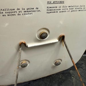 Puede incluir: Primer plano de un objeto circular de color crema con un soporte met&aacute;lico. El objeto tiene un peque&ntilde;o bot&oacute;n y dos remaches. El soporte es de metal oxidado. Texto en varios idiomas es visible en la superficie del objeto.