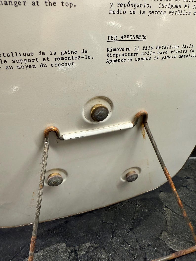 Puede incluir: Primer plano de un objeto circular de color crema con un soporte met&aacute;lico. El objeto tiene un peque&ntilde;o bot&oacute;n y dos remaches. El soporte es de metal oxidado. Texto en varios idiomas es visible en la superficie del objeto.