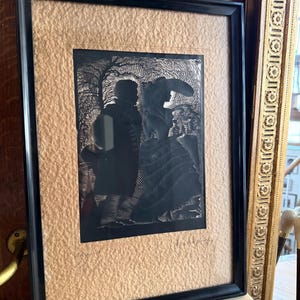 Könnte beinhalten: Gerahmtes schwarzes Silhouette-Kunstwerk, das zwei Figuren in einer Landschaft zeigt. Das Kunstwerk ist vor einem strukturierten, beigefarbenen Hintergrund platziert und in einem schwarzen Holzrahmen gerahmt. Das Kunstwerk wird auf einer Marmoroberfläche ausgestellt.