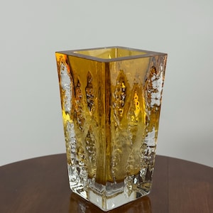 Könnte beinhalten: Eine quadratische Glasvase mit einem leuchtend bernsteinfarbenen Kern, der in klares, strukturiertes Glas an der Außenseite übergeht. Die Oberfläche zeigt ein sich wiederholendes vertikales Muster aus erhabenen, organischen, eisartigen Kristallstrukturen.