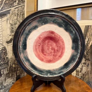 Könnte beinhalten: Ein Keramikteller mit dunklem Rand, einem tealfarbenen Band und einem roten Zentrum. Der Teller wird von einem schwarzen Ständer getragen und steht auf einem Holztisch. Das Design des Tellers erinnert an ein Ziel.