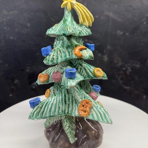 Könnte beinhalten: Eine Keramik-Weihnachtsbaumfigur mit Stern und Kometendetail an der Spitze. Der grüne Baum ist mit bunten Ornamenten geschmückt. Der Sockel ist braun und der Baum steht auf einer weißen Oberfläche.