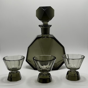 Può includere: Una caraffa in vetro color fumo con un tappo geometrico sfaccettato e un set coordinato di tre piccoli bicchieri a bordo largo. Ogni pezzo presenta una base pesante a più lati e un distinto design angolare realizzato in vetro grigio scuro.