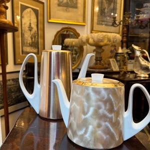 May include: Two vintage teapots with white handles and spouts. One is silver with vertical lines, and the other is gold with a swirling pattern. Both have white lids and sit on a dark wooden surface. The background includes framed artwork.