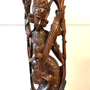 Può includere: Scultura in legno dettagliata di una figura con copricapo e abiti ornati, che tiene uno strumento musicale. La figura è seduta su un cigno, con intagli intricati. La scultura è di un ricco colore marrone.