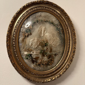 Op de afbeelding: Een antiek, ovaalvormig ingelijst kunstwerk met een goudkleurige, sierlijke lijst. Het kunstwerk binnenin heeft een wit kanten ontwerp met bloemen en groene accenten. Een kleine banner met tekst is zichtbaar.