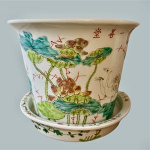 Könnte beinhalten: Weißer Keramik-Blumentopf mit passender Untertasse. Der Topf hat ein handgemaltes Design aus Lotusblumen und Blättern in Grün, Braun und Gelb. Der Topf hat chinesische Schriftzeichen in Rot und ein Vogelpaar.