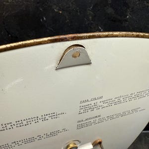 Puede incluir: Primer plano de un objeto circular blanco con un borde dorado. El objeto tiene una percha met&aacute;lica y texto en ingl&eacute;s y espa&ntilde;ol. El texto describe c&oacute;mo colgar el objeto.