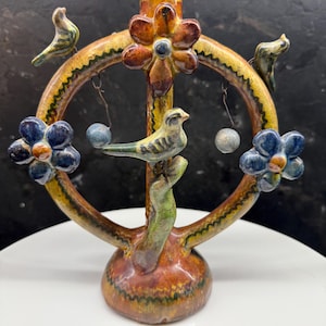 Könnte beinhalten: Ein handgefertigter Kerzenhalter aus Keramik mit einzigartigem Design. Der Halter hat eine zentrale Säule mit einer Blume an der Spitze, einen kreisförmigen Rahmen und dekorative Elemente wie Vögel und Blumen. Die Farbpalette umfasst Braun-, Gelb-, Blau- und Grüntöne.