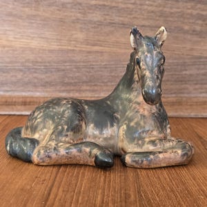 May include: A ceramic figurine of a resting horse. The horse is primarily dark gray with lighter brown and tan markings. The figurine is posed lying down with its legs tucked beneath its body. The background is a wooden surface.