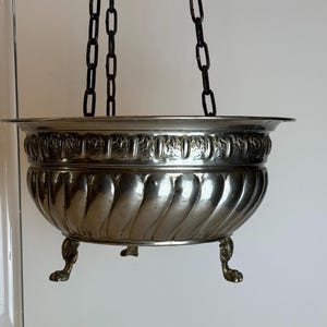 Puede incluir: Un cuenco colgante de metal plateado con un borde ancho y acampanado, y un cuerpo profundo y redondeado con detalles estriados verticales. La sección superior tiene una banda floral en relieve y la base descansa sobre tres patas de garra.