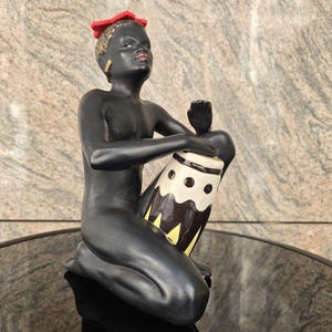 Könnte beinhalten: Eine schwarze Keramikfigur einer knienden Person, die eine Trommel spielt. Die Figur hat eine rote Kopfbedeckung und goldene Ohrringe. Die Trommel ist weiß, braun und gelb.