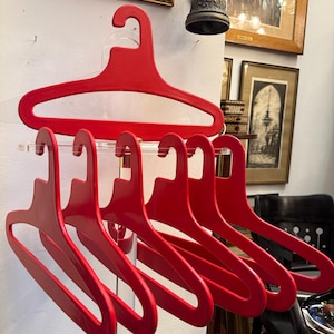 Könnte beinhalten: Ein Set aus sechs roten Kleiderbügeln aus Kunststoff. Ein Bügel wird oben angezeigt, mit fünf weiteren darunter. Die Bügel befinden sich auf einem transparenten Acryl-Präsentationsständer. Der Hintergrund umfasst gerahmte Kunstwerke und Dekorationsgegenstände.