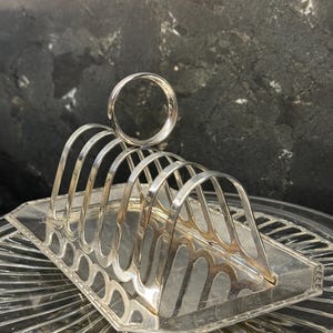 May include: A silver-colored toast rack with arched dividers and a circular handle. The rack sits on a clear, patterned glass plate. The design is classic and elegant, suitable for serving toast or bread.