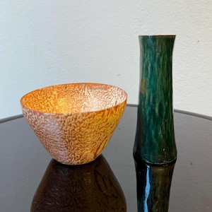 Peut inclure: Un petit bol tacheté orange et marron et un grand vase vert. Le bol a une forme arrondie et le vase est cylindrique. Les deux articles sont sur une surface noire réfléchissante.