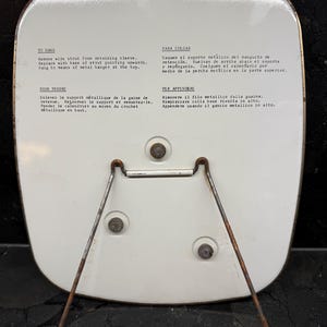 Puede incluir: Un objeto met&aacute;lico rectangular blanco con un soporte met&aacute;lico oxidado y un peque&ntilde;o lazo met&aacute;lico para colgar. La parte posterior del objeto tiene texto en varios idiomas, incluidos ingl&eacute;s, franc&eacute;s, espa&ntilde;ol e italiano.