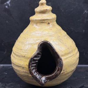 Könnte beinhalten: Ein handgefertigtes, gelb getöntes Keramik-Vogelhaus mit einer dunkelbraunen, tropfenförmigen Öffnung. Das Vogelhaus hat eine strukturierte Oberfläche und eine dekorative, konische Oberseite. Die Gesamtform ist abgerundet und bauchig.