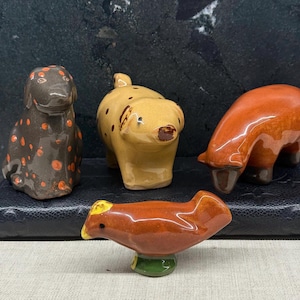 Könnte beinhalten: Vier Keramik-Tierfiguren: ein Hund mit orangefarbenen Flecken, ein gelbes Schwein mit braunen Flecken, ein braunes Schwein und ein brauner Vogel mit gelbem Schnabel. Die Figuren sind glasiert und haben ein glänzendes Finish.