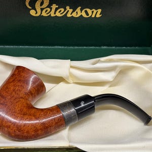 May include: A Peterson tobacco pipe with a curved stem, resting in a green box with the brand name "Peterson" in gold lettering. The pipe is brown with a silver band and black mouthpiece, set against a cream-colored fabric.