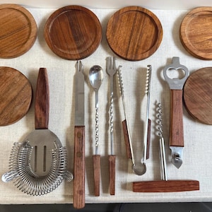 Könnte beinhalten: Eine Sammlung von Barwerkzeugen und Holzuntersetzern. Die Barwerkzeuge umfassen einen Cocktail-Strainer, ein Messer, einen Löffel, eine Gabel, eine Zange, einen Flaschenöffner und einen Korkenzieher. Die Werkzeuge haben Holzgriffe und Edelstahlkomponenten. Die Untersetzer sind rund und aus Holz.