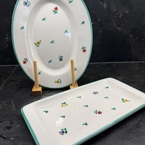 Könnte beinhalten: Zwei weiße Keramik-Servierplatten mit floralem Design. Die ovale Platte steht auf einem Holzständer. Beide Platten haben einen grünen Rand und sind mit kleinen bunten Blumen und grünen Blättern verziert.
