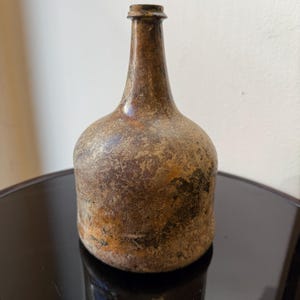 May include: An antique brown bottle with a rounded base and a long, narrow neck. The bottle has a textured surface with areas of discoloration, indicating age and use. The bottle is displayed on a dark, reflective surface.
