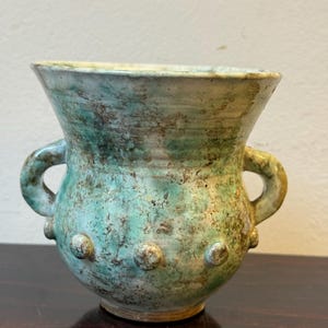 Könnte beinhalten: Eine handgefertigte Keramikvase mit strukturierter Oberfläche und einem einzigartigen, gealterten Aussehen. Die Vase hat eine türkisfarbene und braune Farbpalette, zwei Griffe und dekorative, erhabene Noppen. Das Gefäß hat eine weite Öffnung und einen runden Boden.